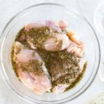 Raw chicken pieces marinating in a clear glass bowl with herbs and seasoning, placed on a light-colored surface.