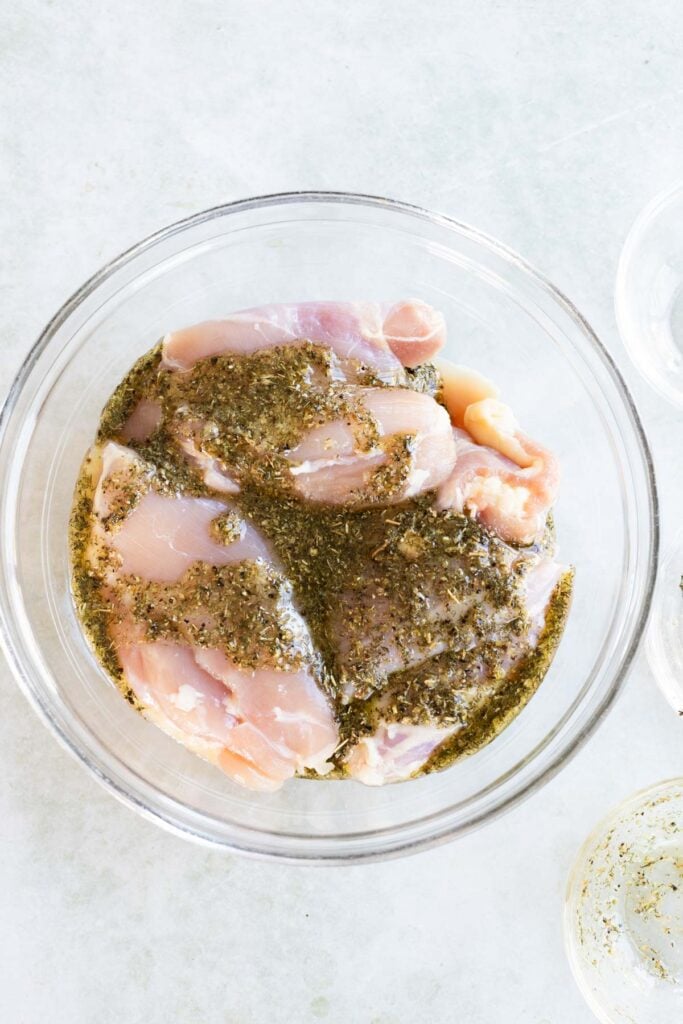 Raw chicken pieces marinating in a clear glass bowl with herbs and seasoning, placed on a light-colored surface.