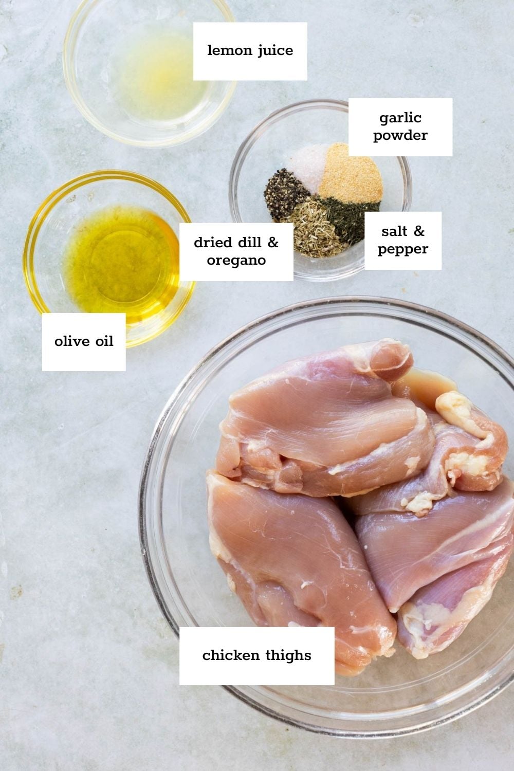 Raw chicken thighs in a glass bowl with small bowls of olive oil, lemon juice, dried dill, oregano, garlic powder, salt, and pepper on a light surface. Labels identify each ingredient.