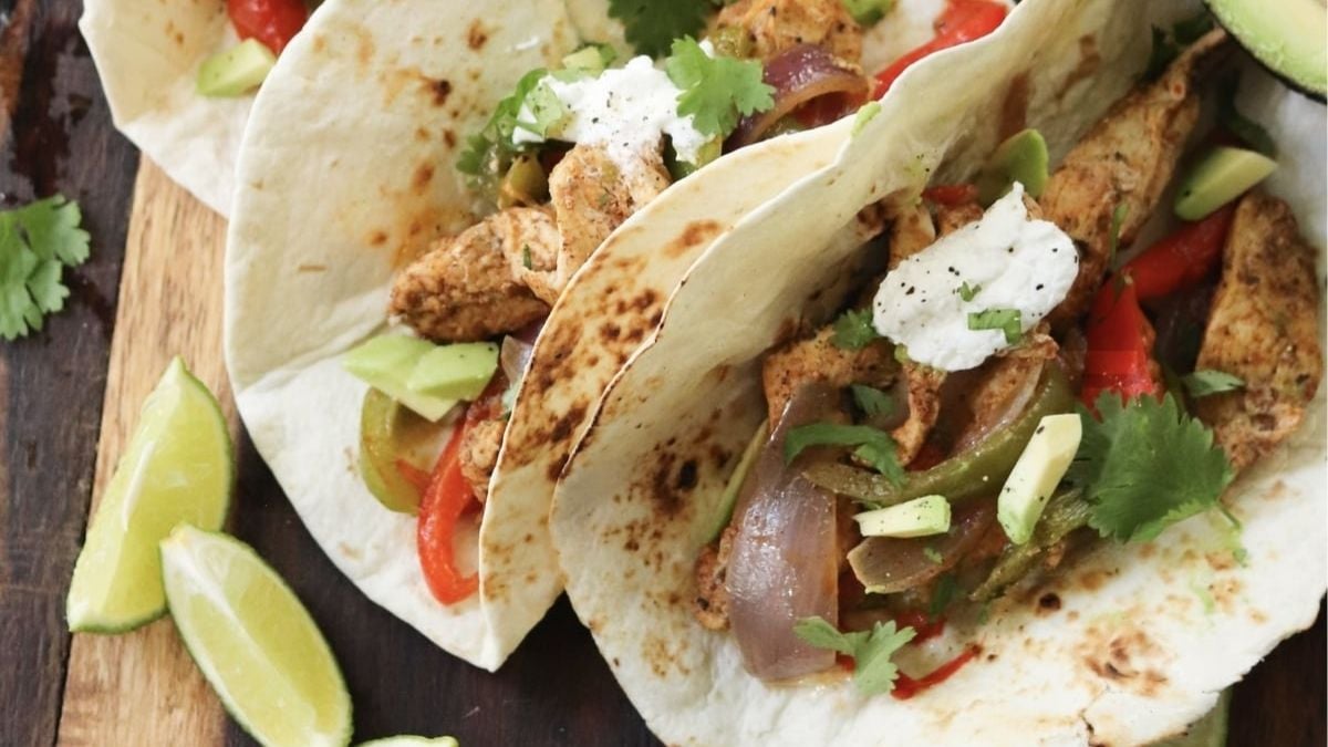 Three chicken fajita tacos with grilled vegetables, avocado slices, cilantro, and a dollop of sour cream on flour tortillas, served with lime wedges on a wooden board.