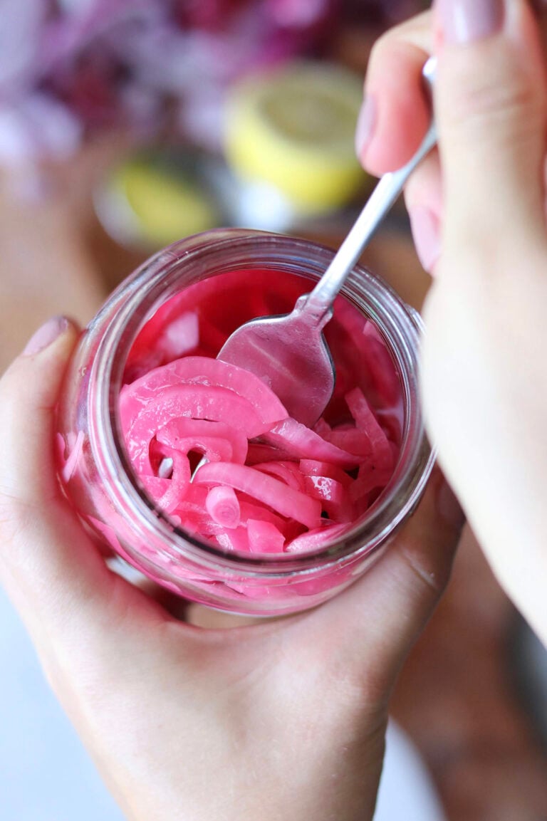 A hand holds a jar of pickled red onions while another hand uses a spoon to lift some of the onions from the jar.
