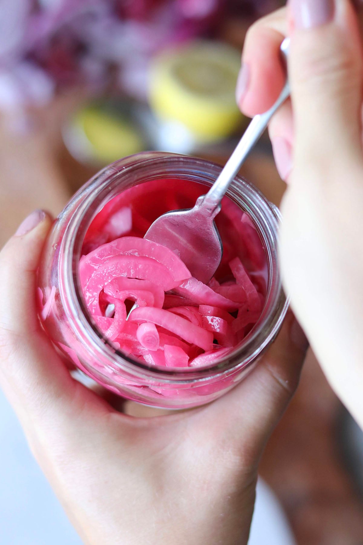 A hand holds a jar of pickled red onion while another hand uses a spoon to lift some of the onions from the jar.