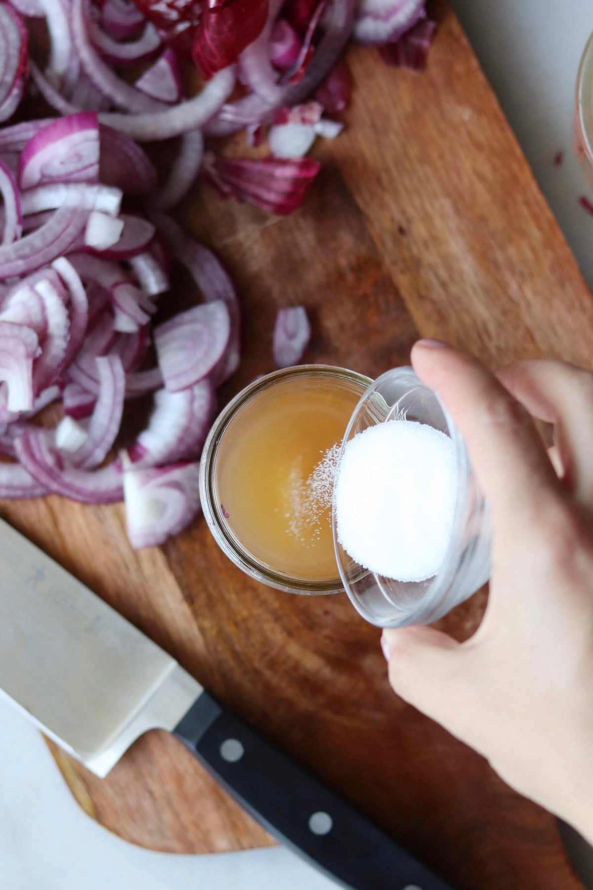 A hand pours salt from a small bowl into a small glass jar of liquid,