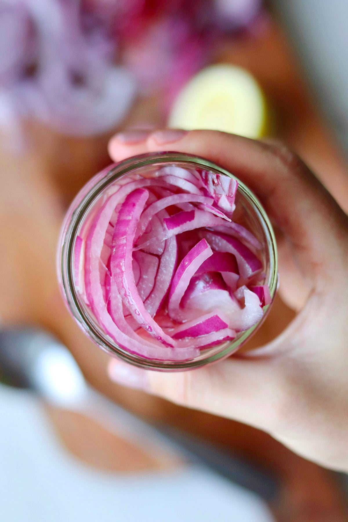 A hand holding a glass jar filled with thin slices of onion, viewed from above.