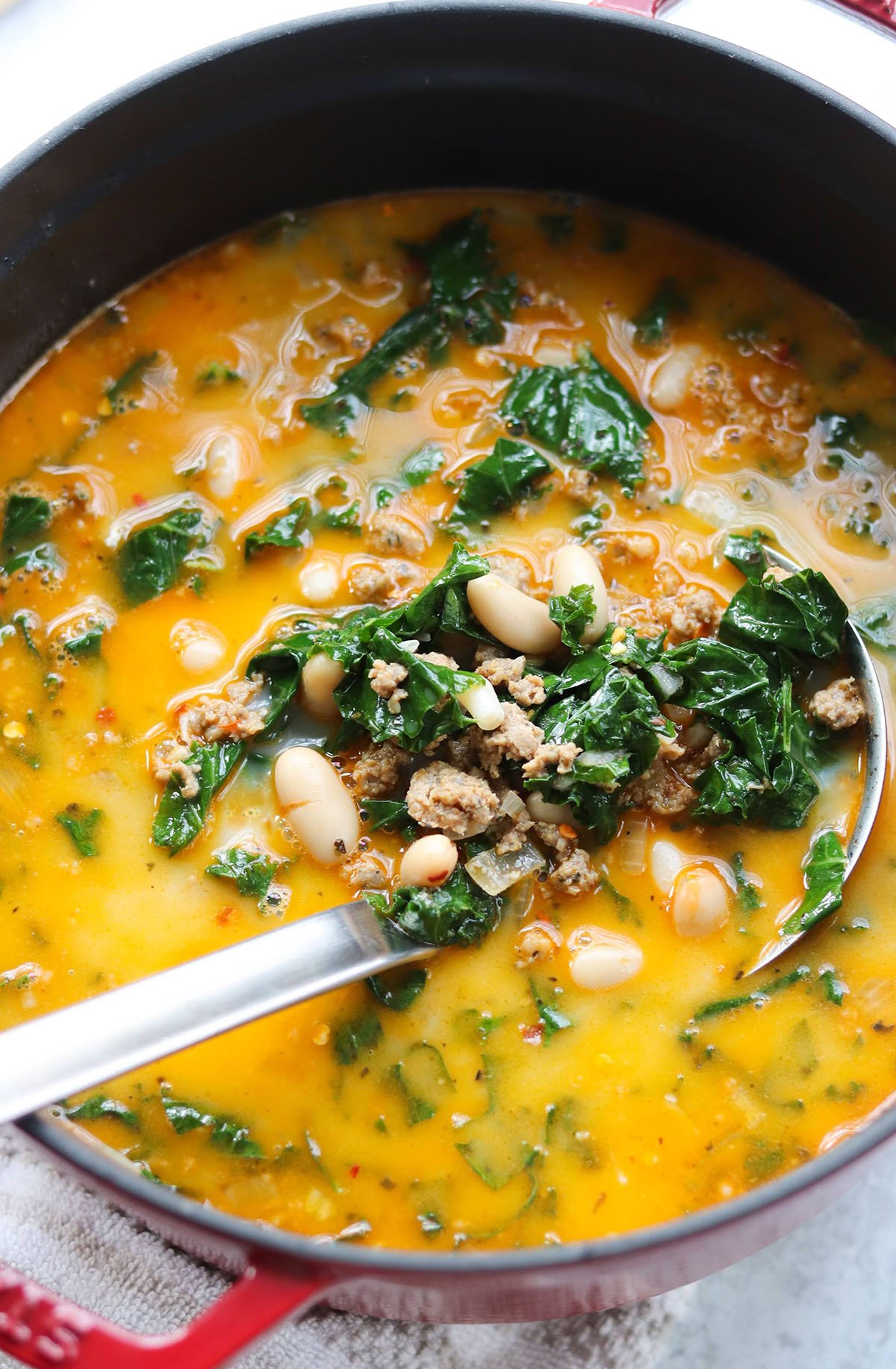 A pot of Tuscan bean soup with leafy greens, ground sausage, and broth, with a ladle resting inside.