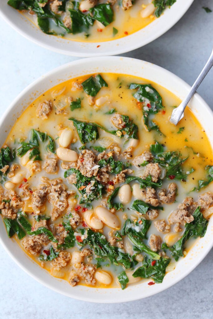 A bowl of soup containing ground meat, white beans, chopped greens, and broth, with a spoon resting in the bowl.