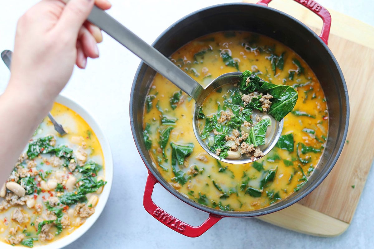 A hand ladles sausage, white bean, and kale soup from a red pot into a bowl on a light countertop.