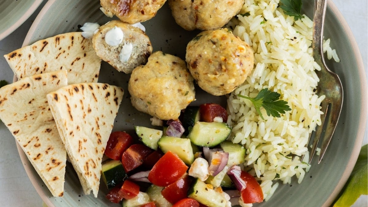 A plate with pita bread slices, chicken meatballs, white rice, and a cucumber-tomato salad with feta cheese, garnished with parsley.