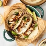 Two fish tacos with grilled fish, shredded cabbage, and cilantro on tortillas, served in a dish with lime wedges, surrounded by tortillas, sauce, and herbs.