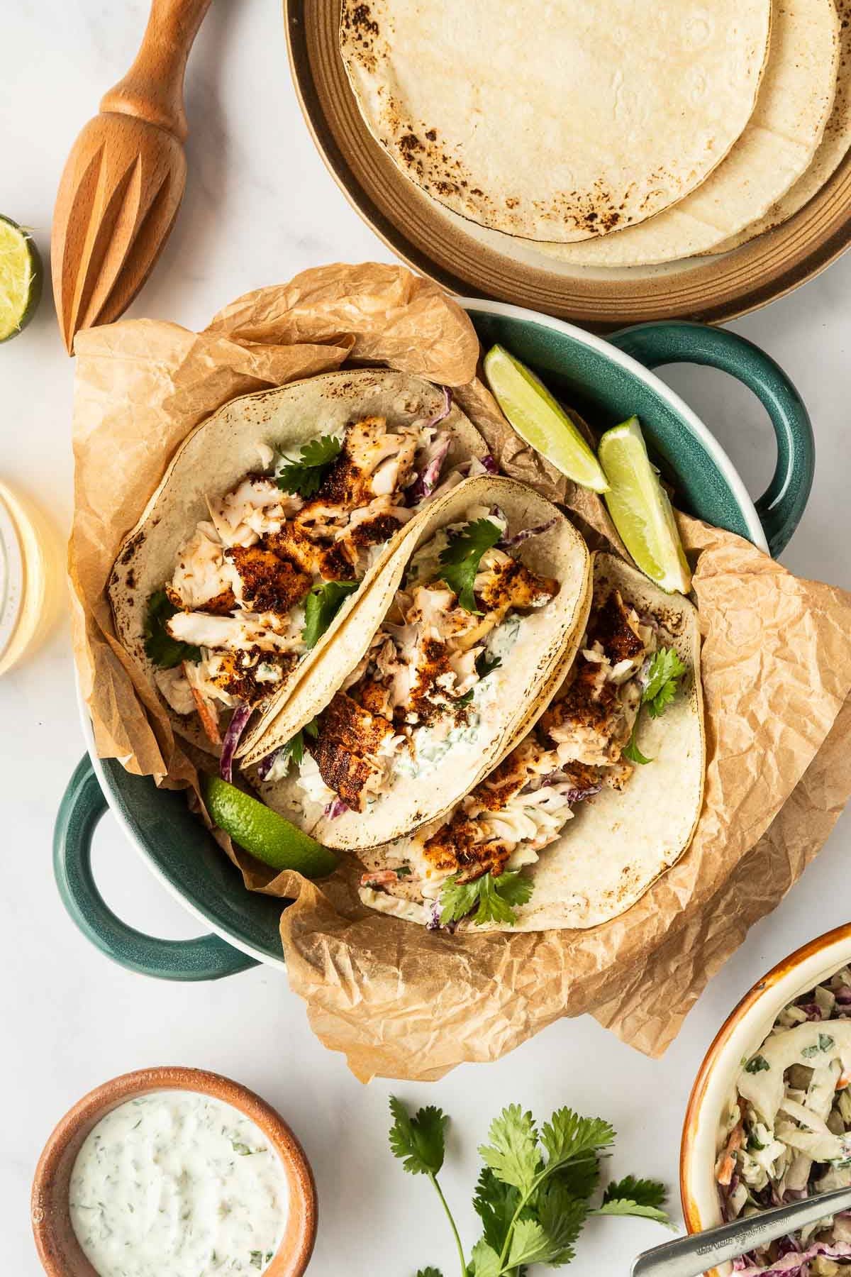 Two fish tacos with grilled fish, shredded cabbage, and cilantro on tortillas, served in a dish with lime wedges, surrounded by tortillas, sauce, and herbs.