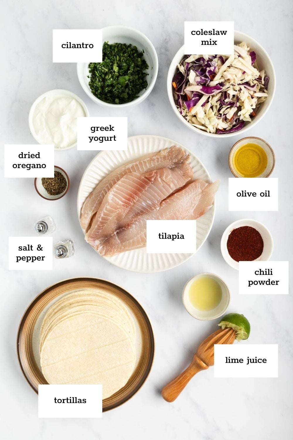 Ingredients for fish tacos laid out on a white surface, including tilapia, tortillas, coleslaw mix, cilantro, greek yogurt, dried oregano, salt, pepper, olive oil, chili powder, and lime juice.