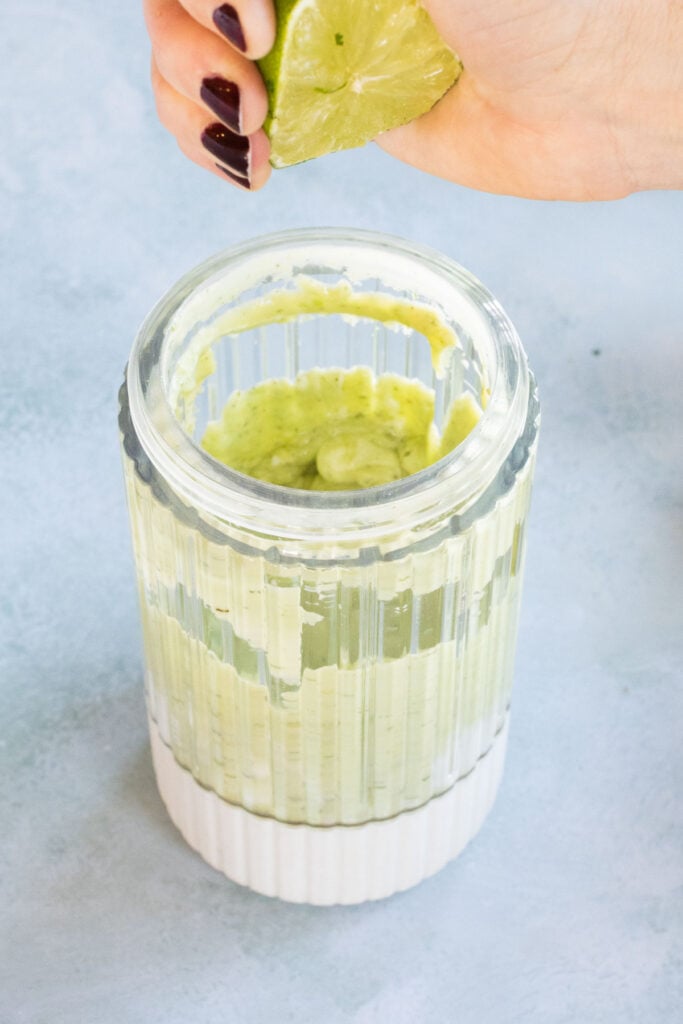 A hand squeezes a lime over a glass container filled with a creamy green dressing on a light blue surface.