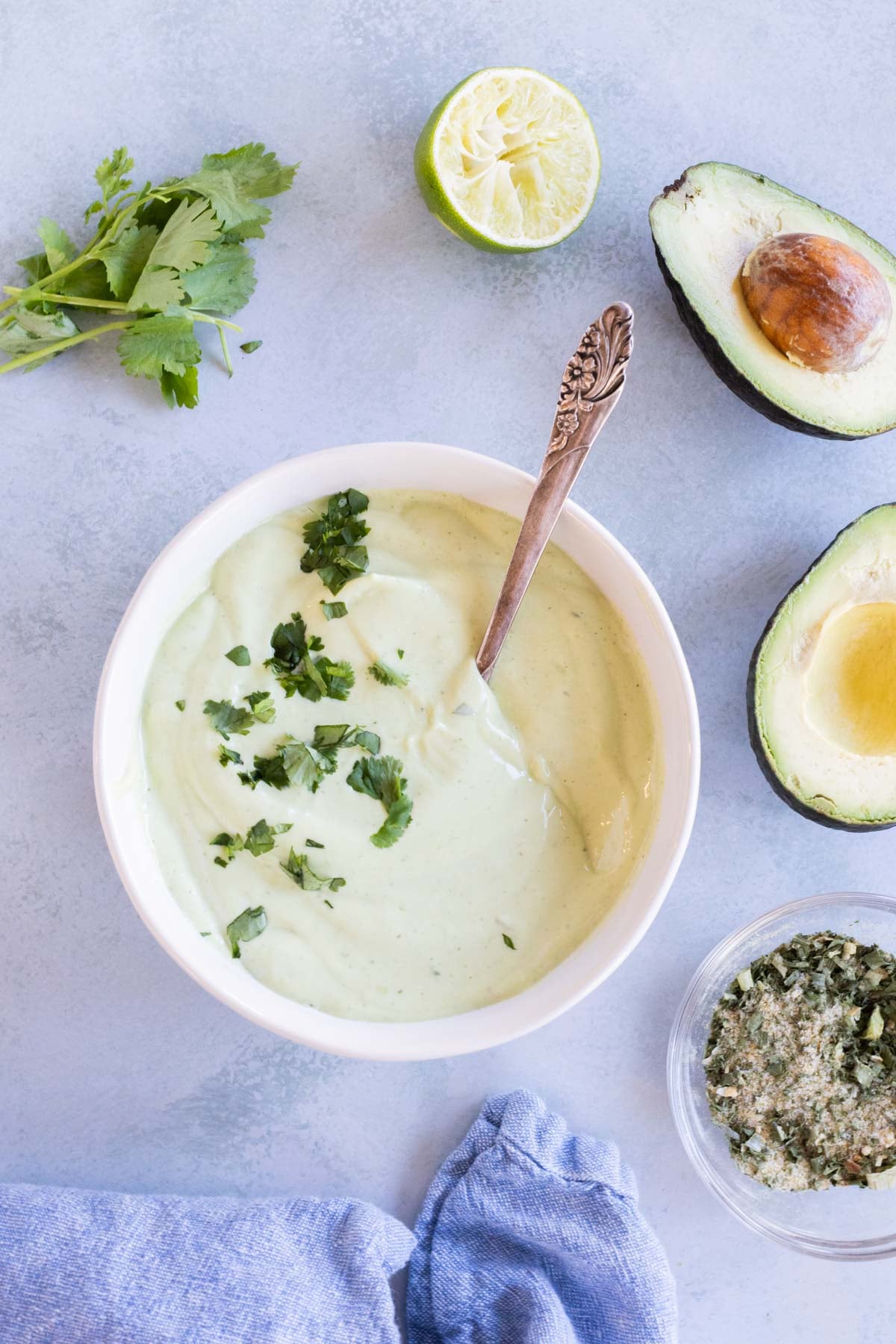 A bowl of avocado lime ranch dressing garnished with chopped herbs sits on a table, surrounded by cilantro, a halved lime, a halved avocado, and a jar of seasoning.
