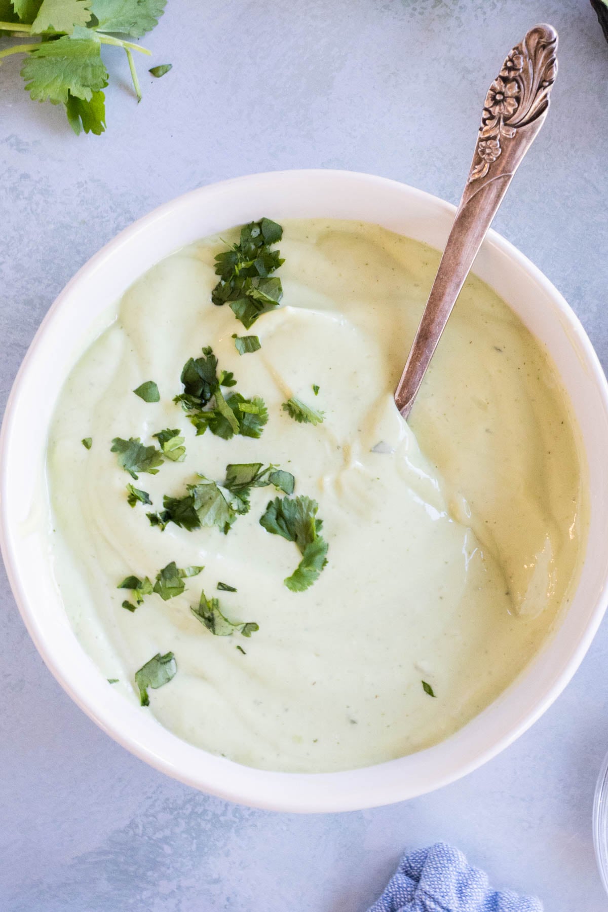 A bowl of avocado lime ranch dressing topped with chopped fresh herbs, with a silver spoon resting inside the bowl, viewed from overhead.