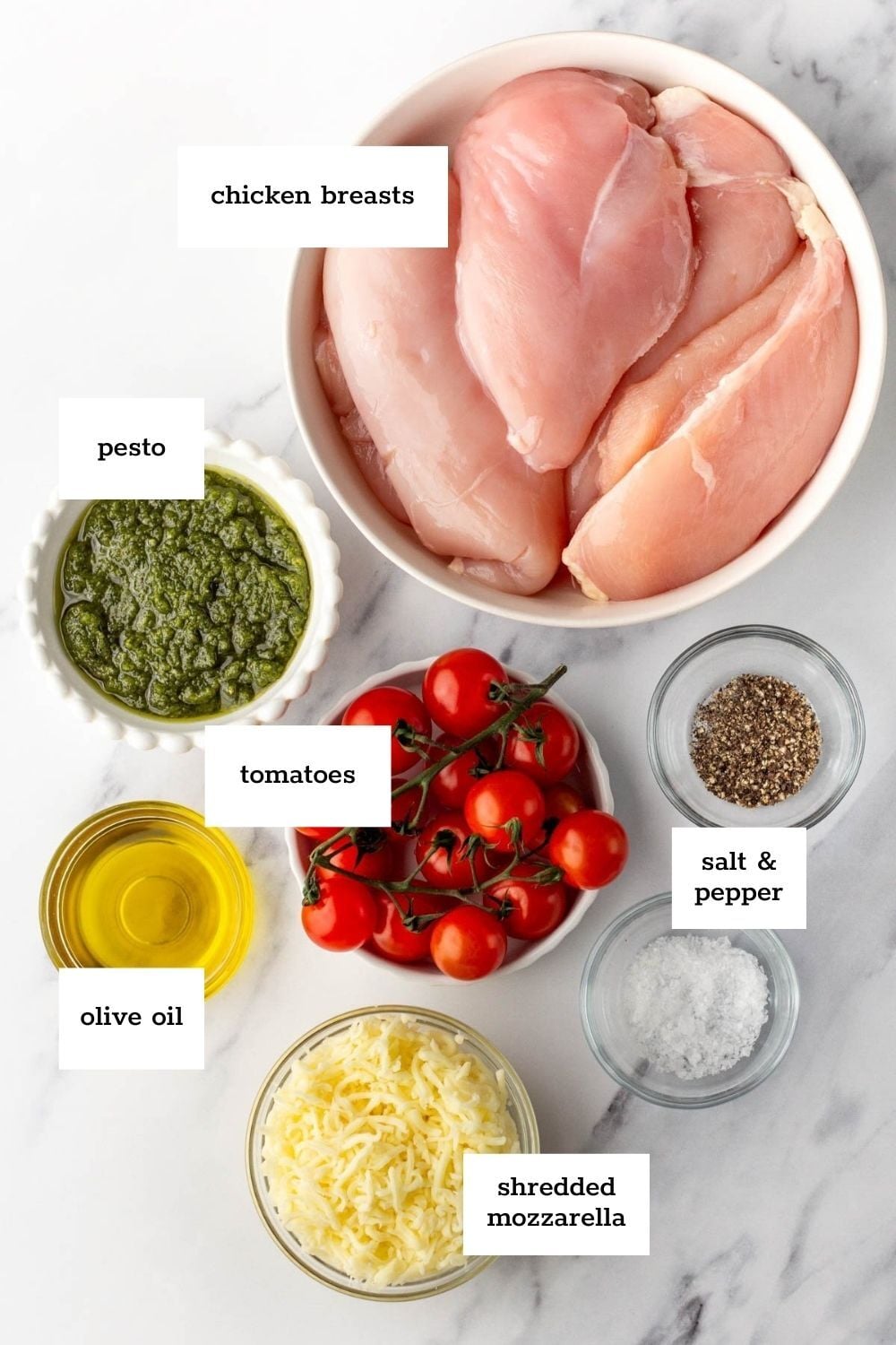 Top-down view of ingredients for a chicken dish: raw chicken breasts, pesto, cherry tomatoes, olive oil, shredded mozzarella, salt, and pepper, each labeled.