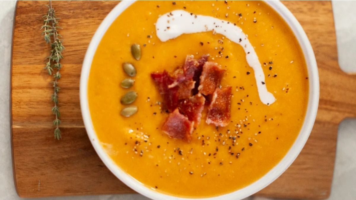 Bowl of orange soup topped with bacon pieces, pumpkin seeds, black pepper, and a swirl of cream, on a wooden board with a sprig of thyme beside it.