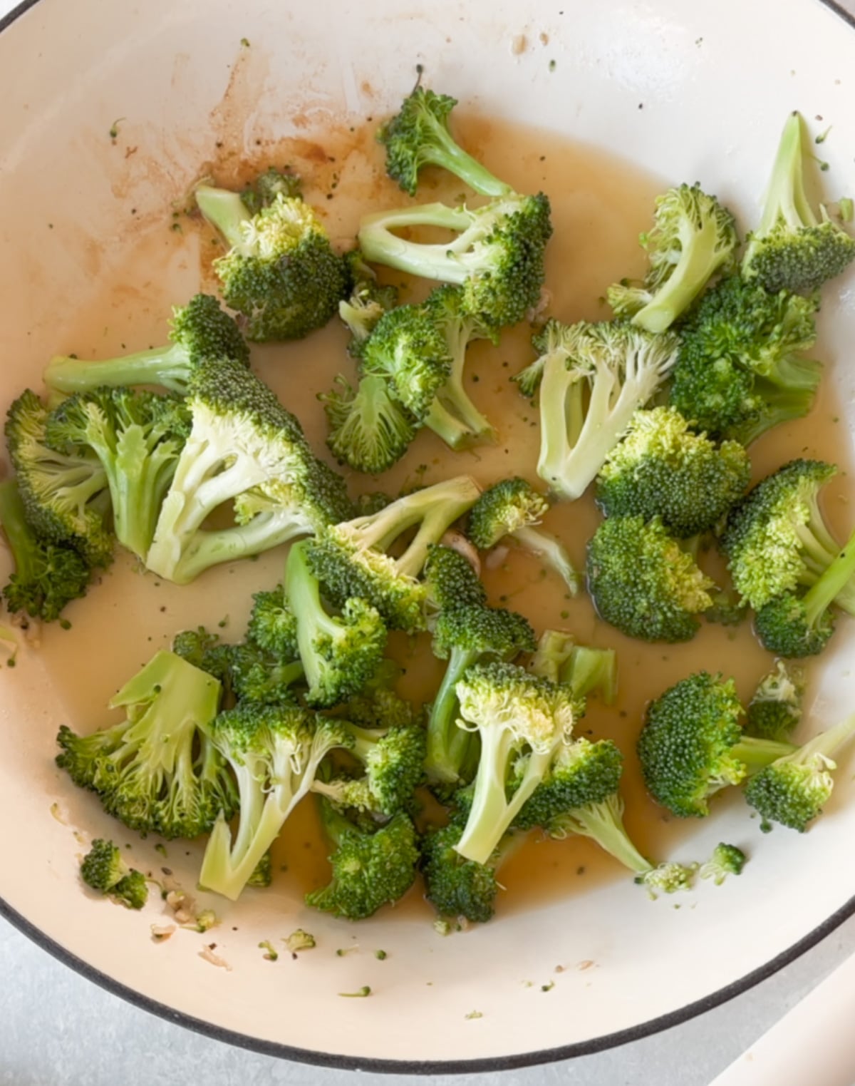 Broccoli florets cooking in a white pan with some liquid and seasonings visible at the bottom.