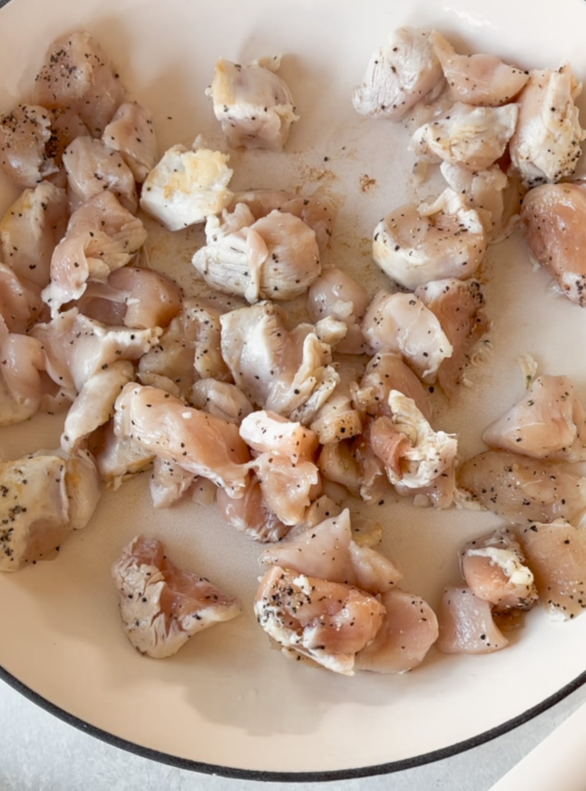 Raw, diced chicken pieces seasoned with black pepper are spread out in a white pan, ready to be cooked.