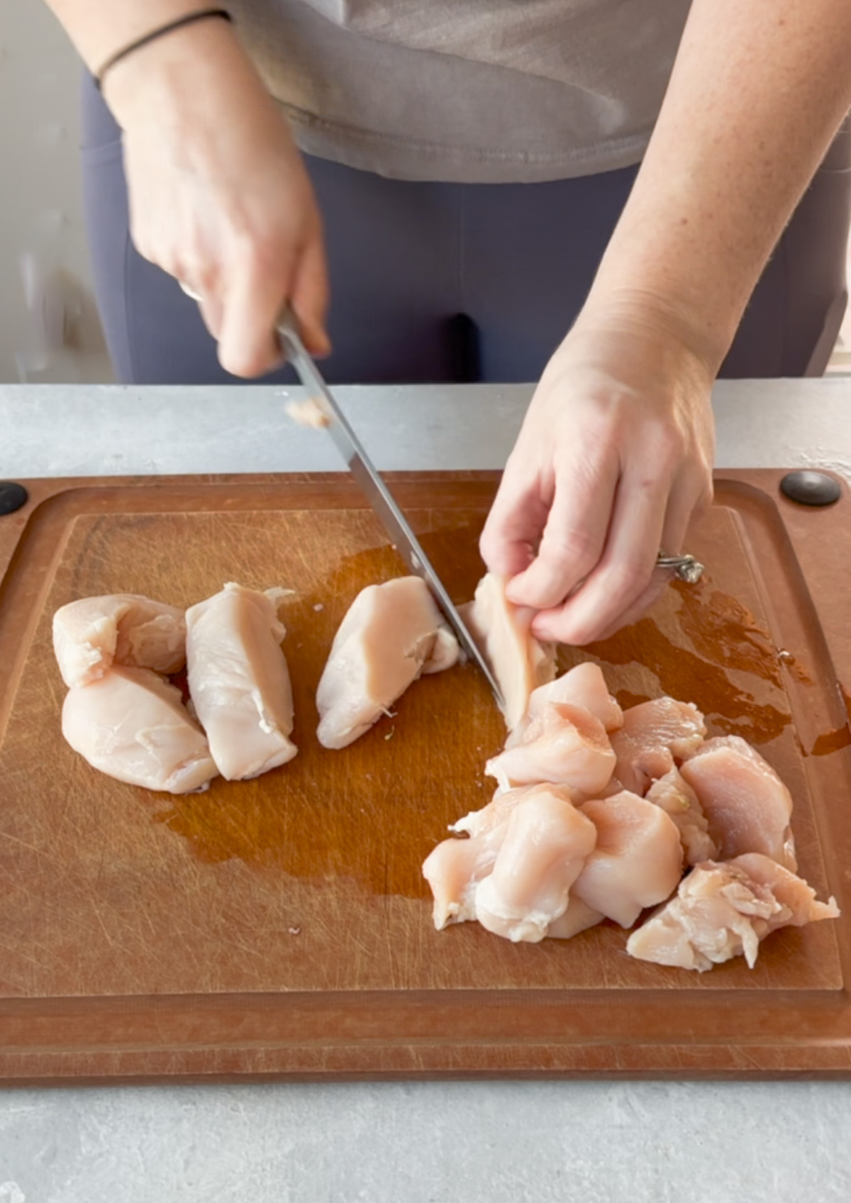A person cuts raw chicken breasts into cubes on a wooden cutting board.