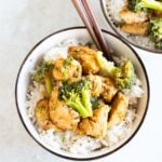 A bowl of white rice topped with cooked chicken pieces and broccoli, with wooden chopsticks resting on the bowl. Another similar bowl is partially visible in the background.