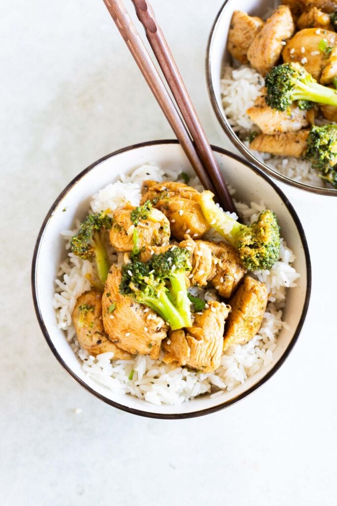 A bowl of white rice topped with cooked chicken pieces and broccoli, with wooden chopsticks resting on the bowl. Another similar bowl is partially visible in the background.