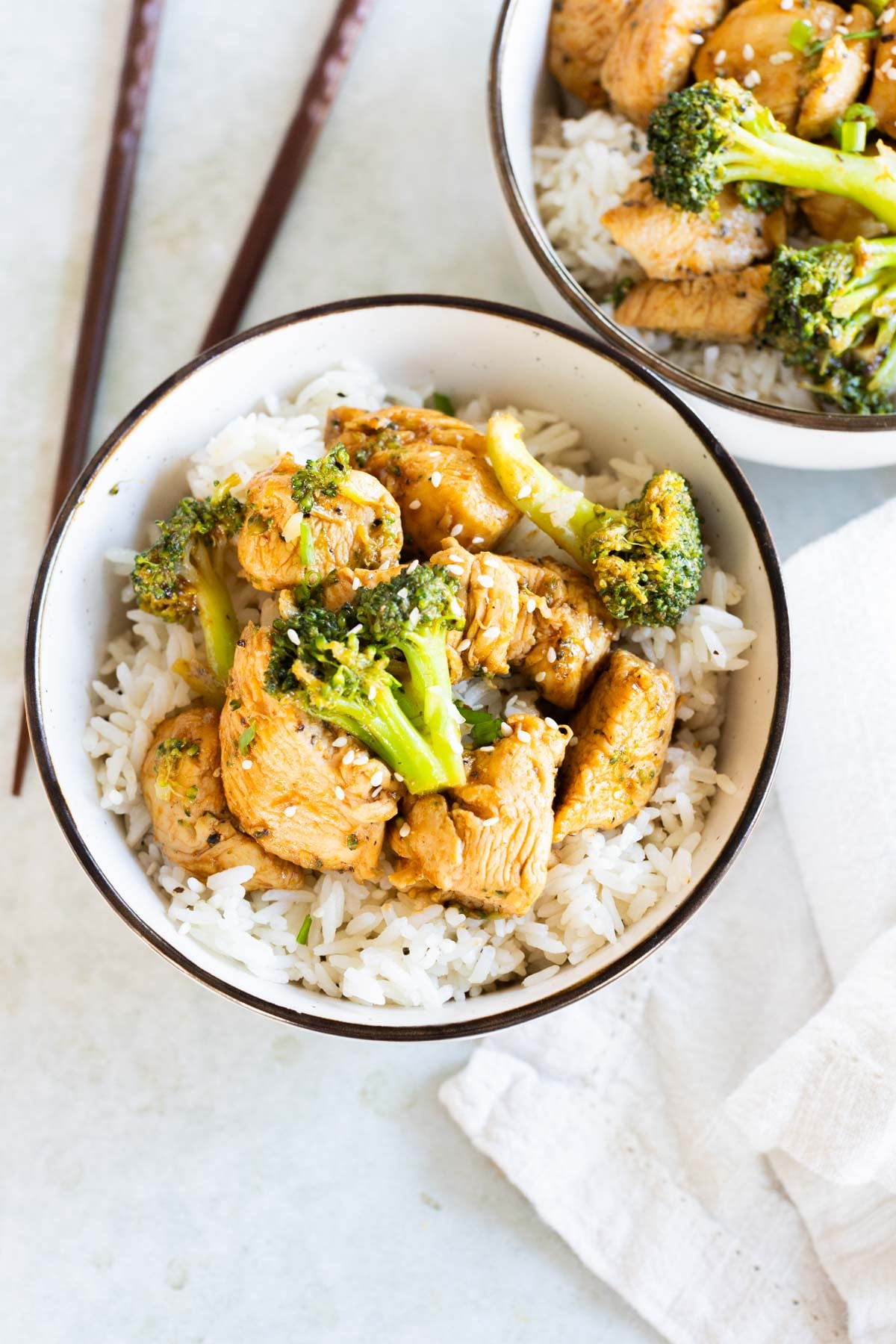 A bowl of white rice topped with cooked chicken pieces, broccoli florets, and sesame seeds. Another similar bowl and chopsticks are nearby.
