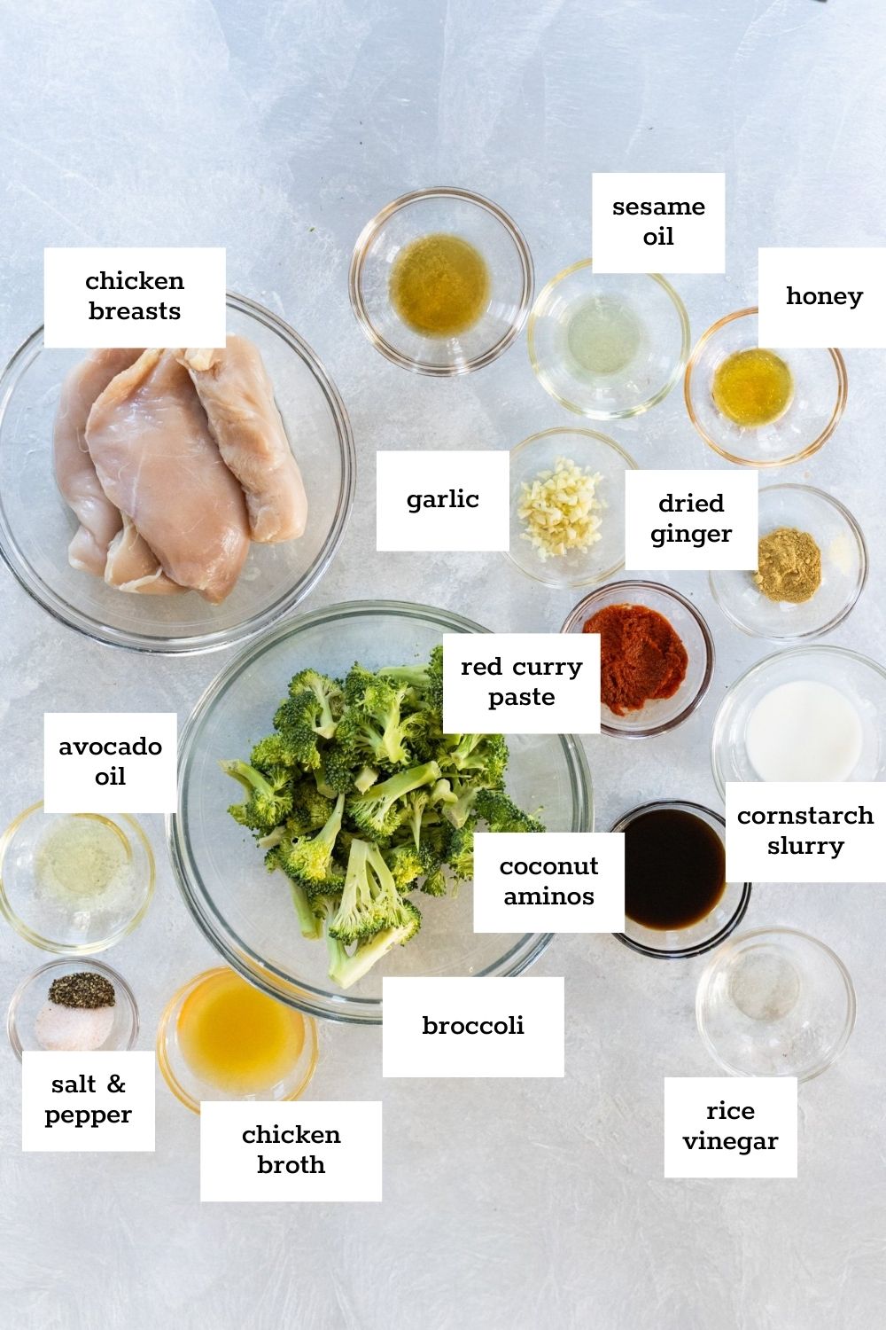 Top-down view of labeled ingredients for a Chinese chicken and broccoli stir fry, including raw chicken breasts, broccoli, oils, seasonings, red curry paste, and various sauces in bowls.