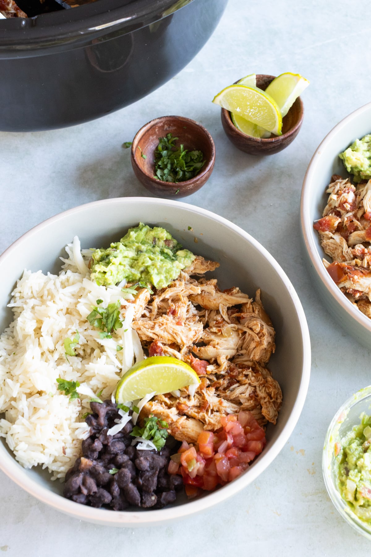 A bowl with rice, shredded chicken, black beans, guacamole, diced tomatoes, and a lime wedge, with small bowls of lime and herbs nearby.