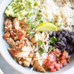 A bowl containing shredded chicken, white rice, black beans, guacamole, pico de gallo, shredded cheese, and a lime wedge, garnished with cilantro.
