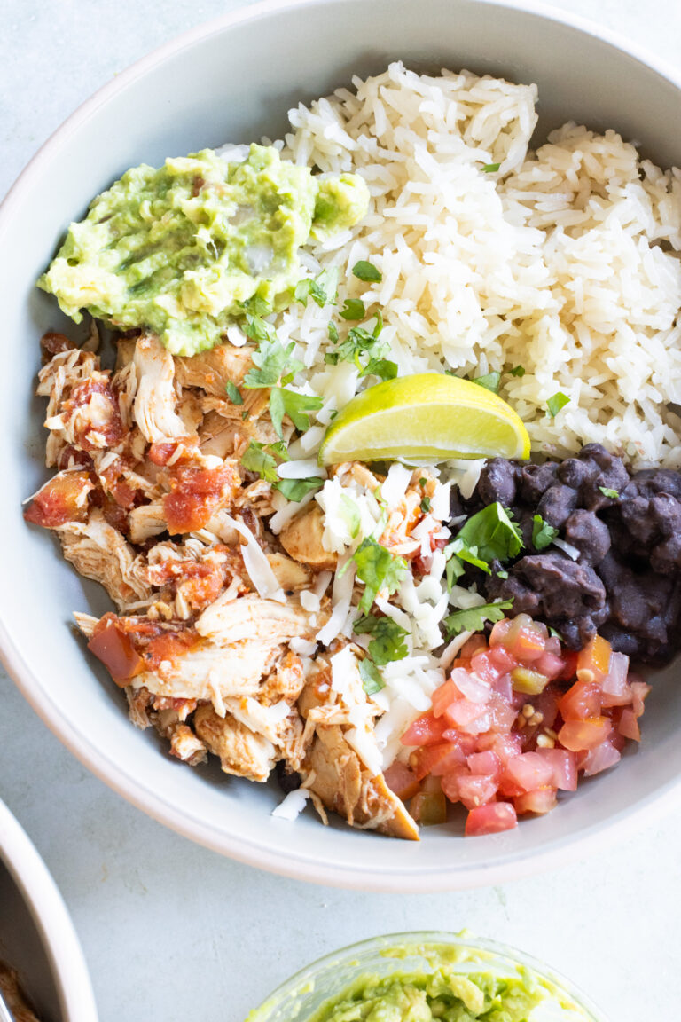 A bowl containing shredded chicken, white rice, black beans, guacamole, pico de gallo, shredded cheese, and a lime wedge, garnished with cilantro.