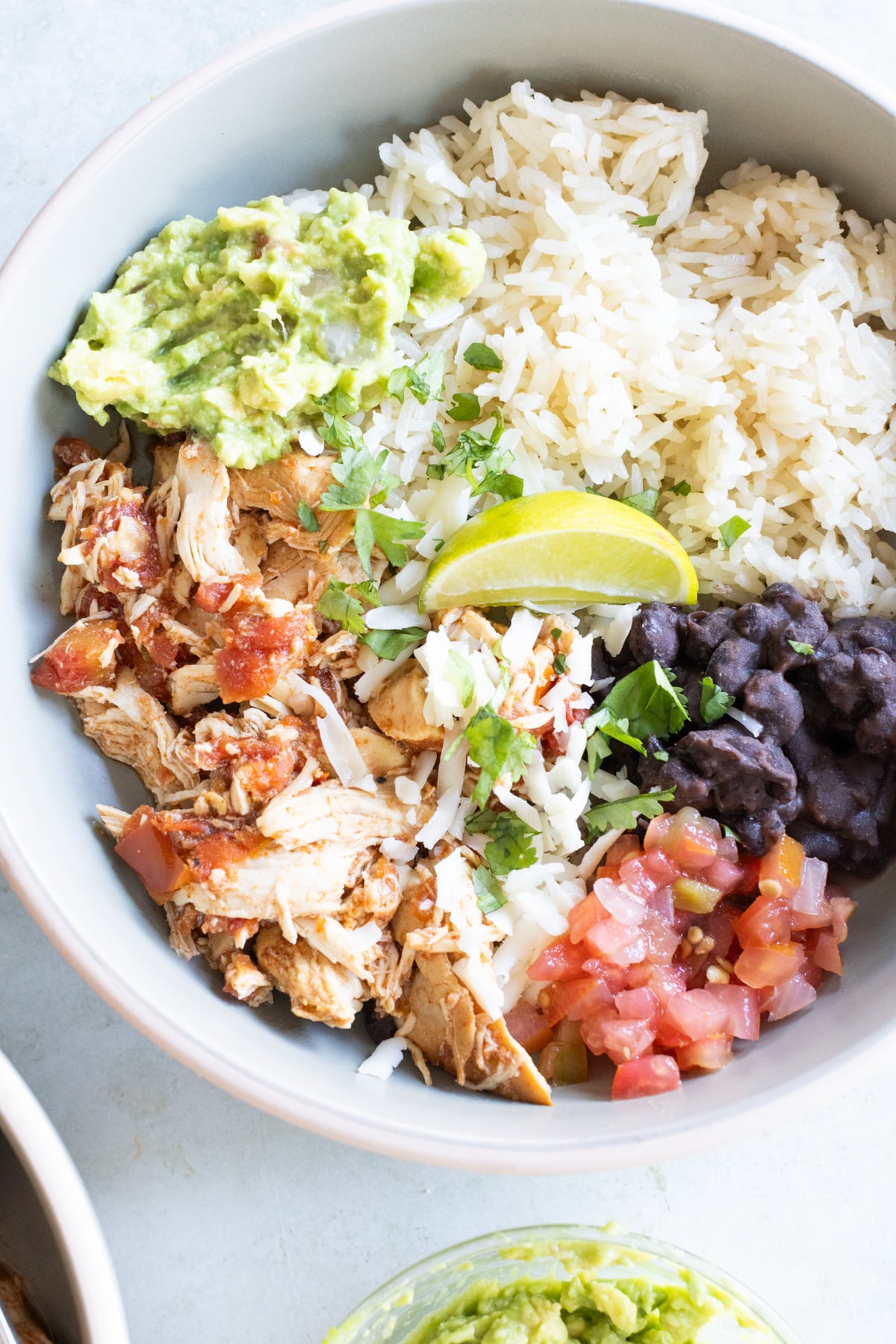 A bowl containing shredded chicken, white rice, black beans, guacamole, pico de gallo, shredded cheese, and a lime wedge, garnished with cilantro.