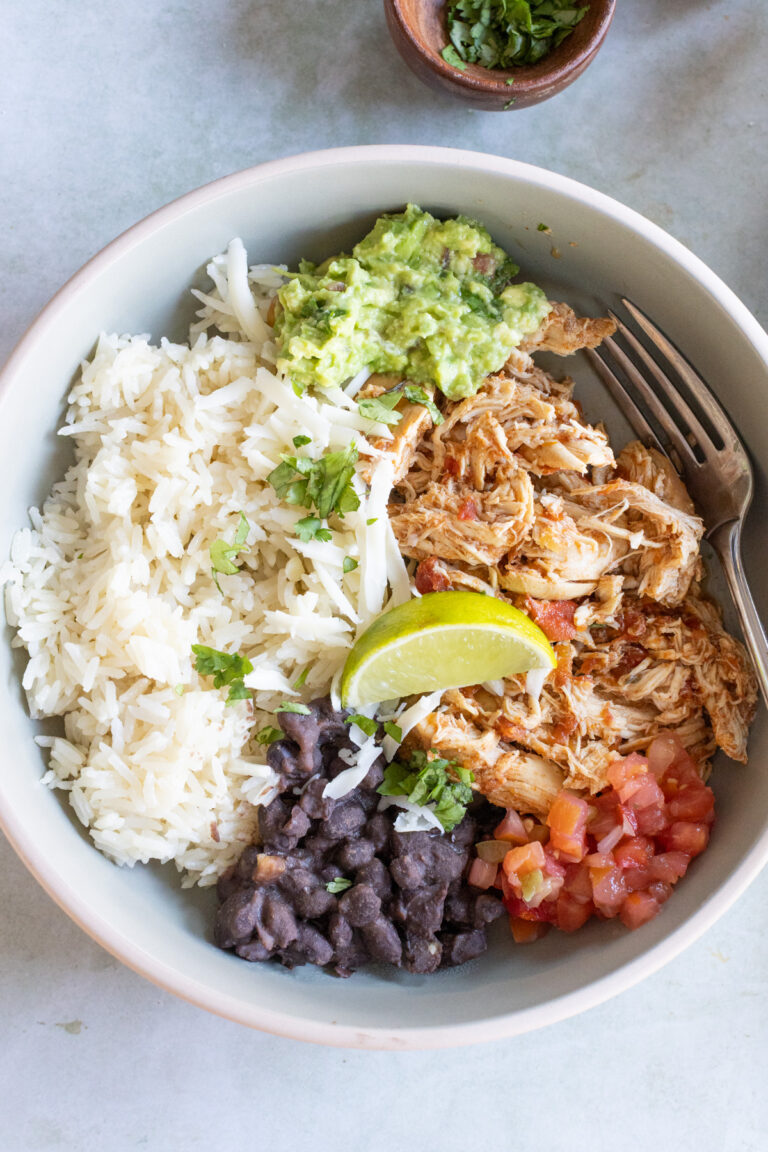 A bowl with white rice, shredded chicken, black beans, pico de gallo, guacamole, a lime wedge, and a fork on the side.