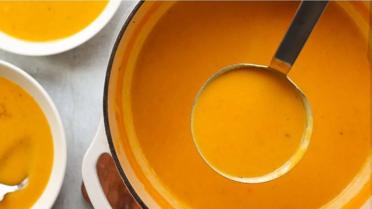 A ladle filled with orange-colored soup hovers over a pot of the same soup, with two bowls of soup nearby on a light surface.