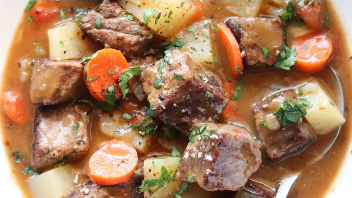 A close-up of beef stew featuring chunks of beef, potato, carrot slices, and parsley in a thick brown broth.