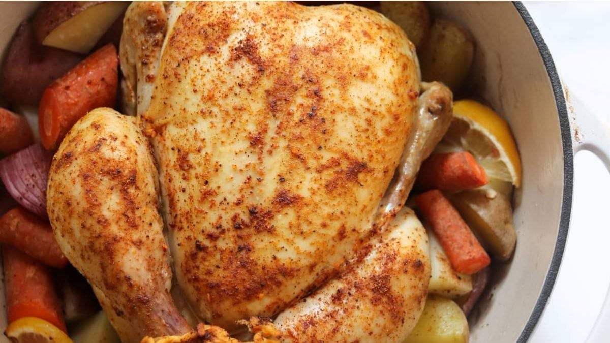 A seasoned whole roasted chicken in a pot, surrounded by carrots, potatoes, red onions, and lemon wedges.