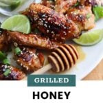 Plate of grilled honey chicken wings garnished with sesame seeds and green onions, served with lime wedges and a honey dipper, next to a bowl of lime slices.