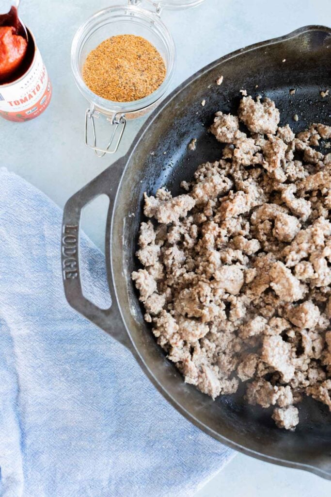 Ground meat cooking in a black cast iron skillet, with a jar of seasoning and a container of tomato paste nearby on a light surface.