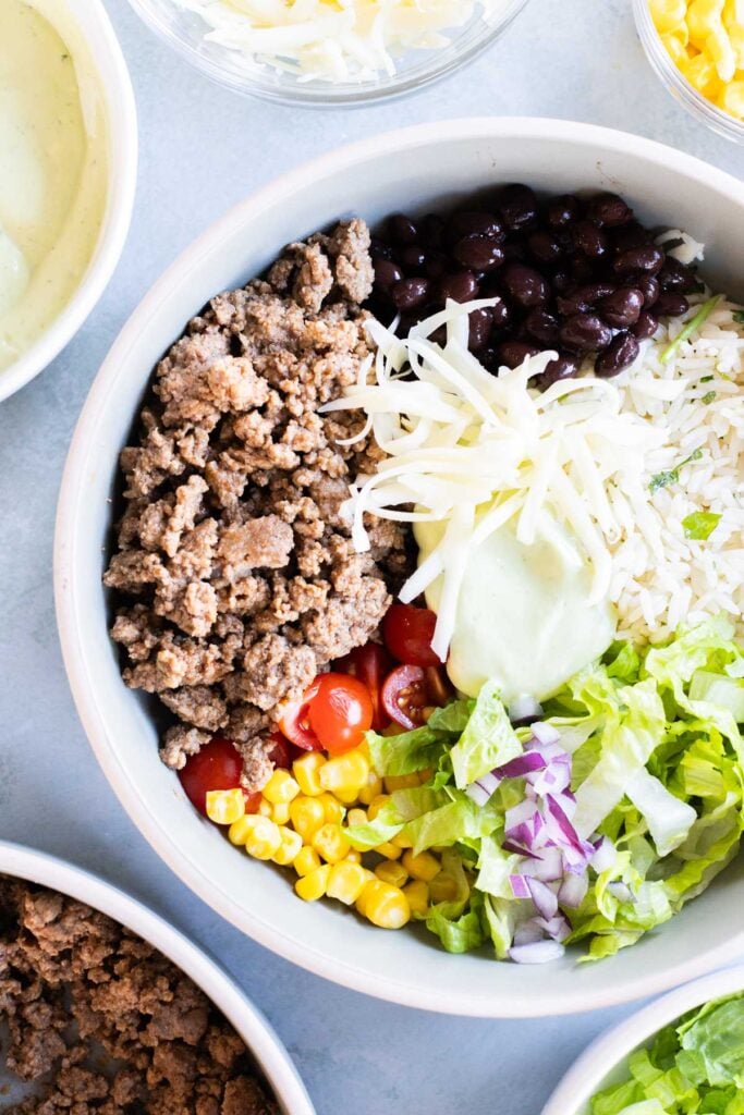 A bowl filled with ground beef, black beans, shredded cheese, rice, cherry tomatoes, corn, lettuce, red onion, and a drizzle of creamy sauce.