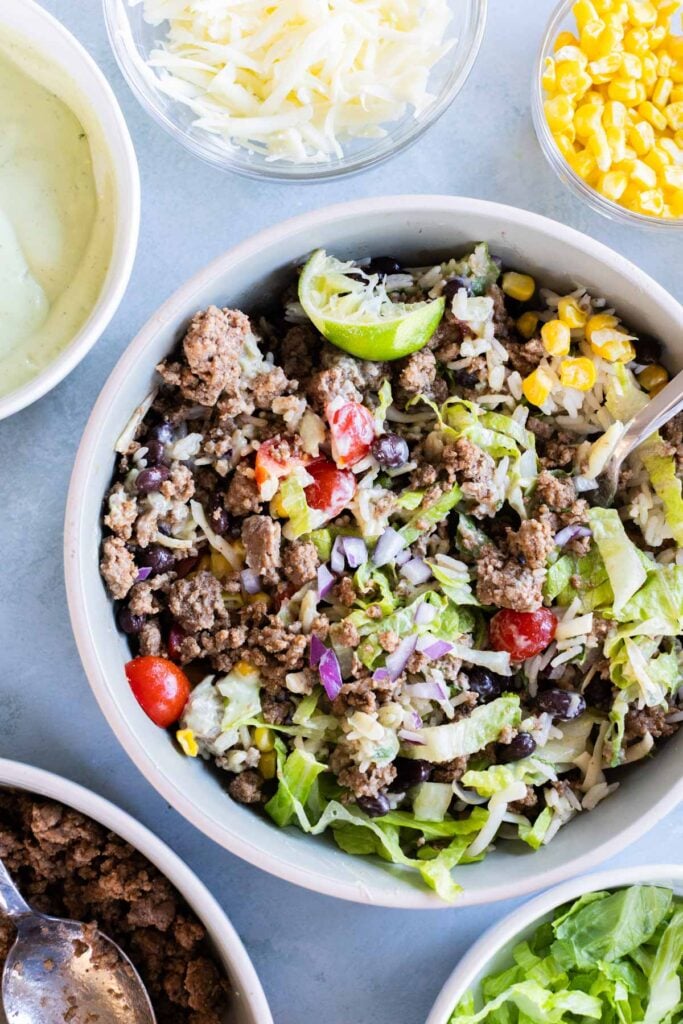 A ground beef burrito bowl with lettuce, tomatoes, corn, black beans, shredded cheese, and a lime wedge, surrounded by bowls of dressing, cheese, corn, and meat.