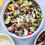 A bowl filled with seasoned ground beef, rice, black beans, corn, lettuce, grape tomatoes, red onion, and avocado dressing, with a lime wedge on the side.