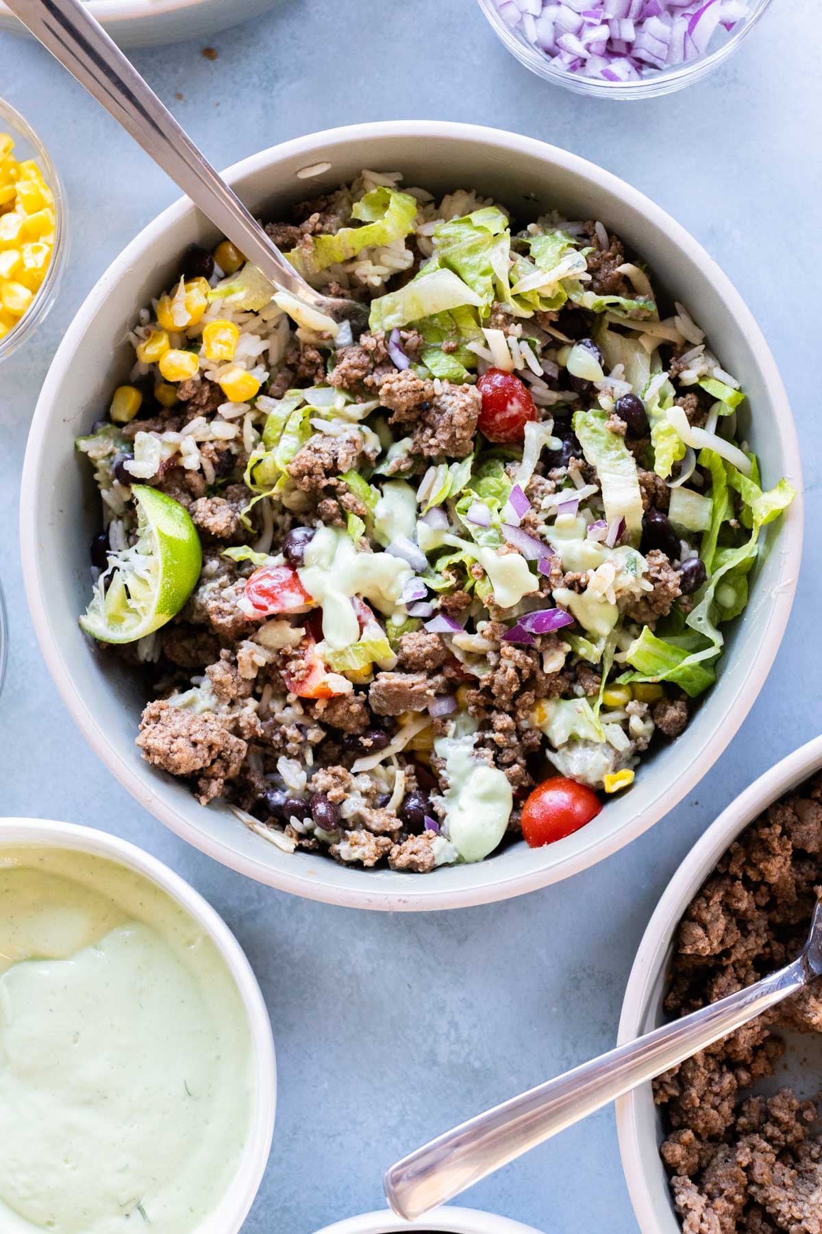 A ground beef burrito bowl filled with seasoned ground beef, rice, black beans, corn, lettuce, grape tomatoes, red onion, and avocado dressing, with a lime wedge on the side.