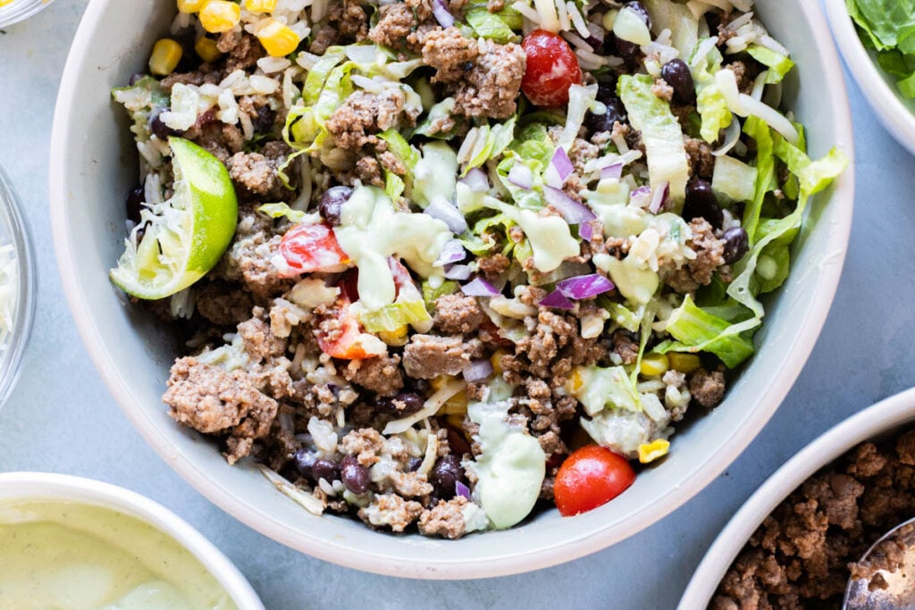 A bowl filled with ground beef, rice, lettuce, black beans, corn, cherry tomatoes, red onions, shredded cheese, a lime wedge, and a drizzle of creamy green sauce.