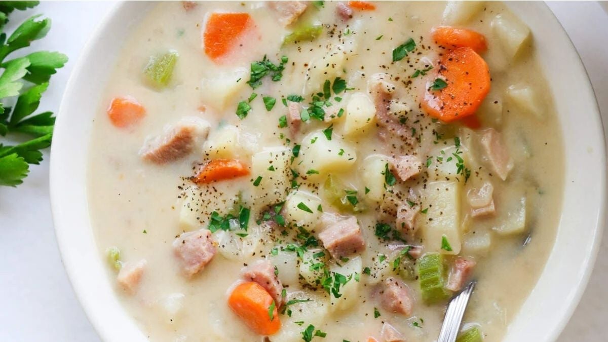 A bowl of creamy potato soup with diced ham, carrots, potatoes, celery, and parsley, topped with black pepper, with a spoon in the bowl.