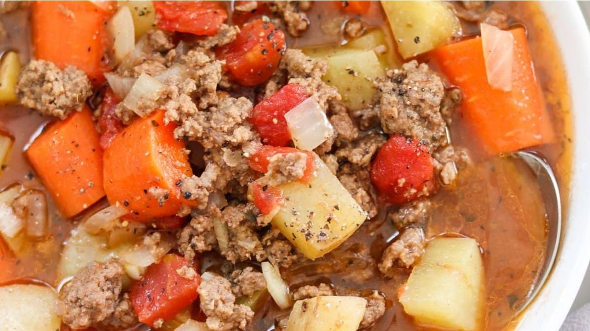 Close-up of a bowl of soup containing ground beef, diced potatoes, carrots, tomatoes, onions, and black pepper in broth.