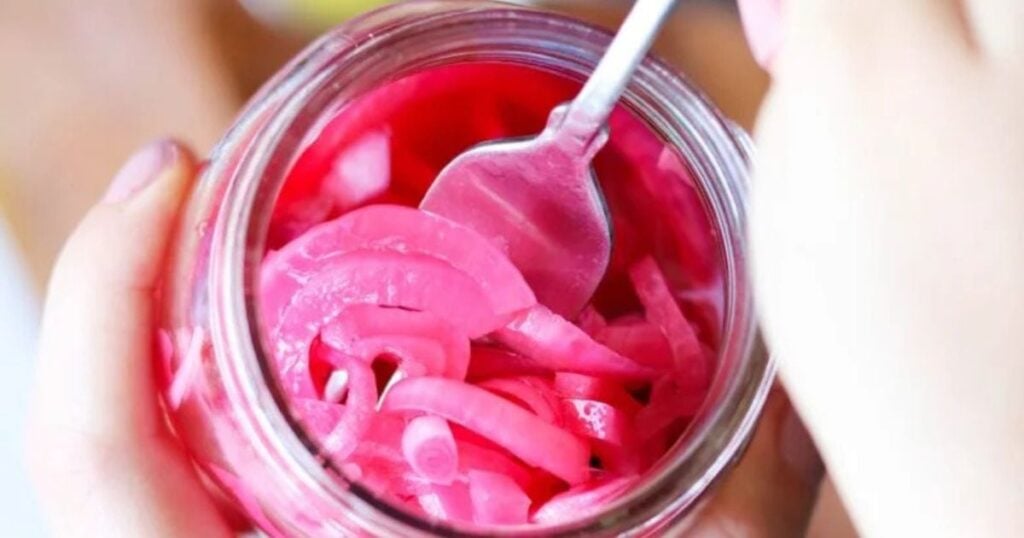 A person holds a glass jar of sliced pickled red onions and uses a spoon to scoop some out.
