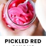 A person holds a jar of sliced pickled red onions and stirs them with a spoon. The label below reads, “PICKLED RED ONIONS cook at home mom.”.