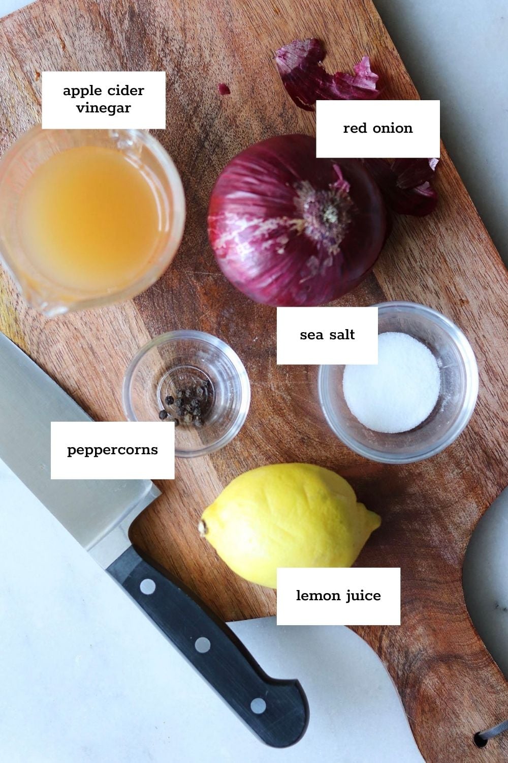Overhead view of labeled ingredients on a cutting board: apple cider vinegar, a red onion, sea salt, lemon, and peppercorns.