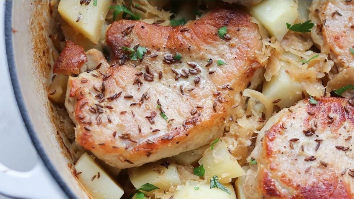Close-up of browned pork chops cooked with potatoes, sauerkraut, caraway seeds, and parsley in a white dish.