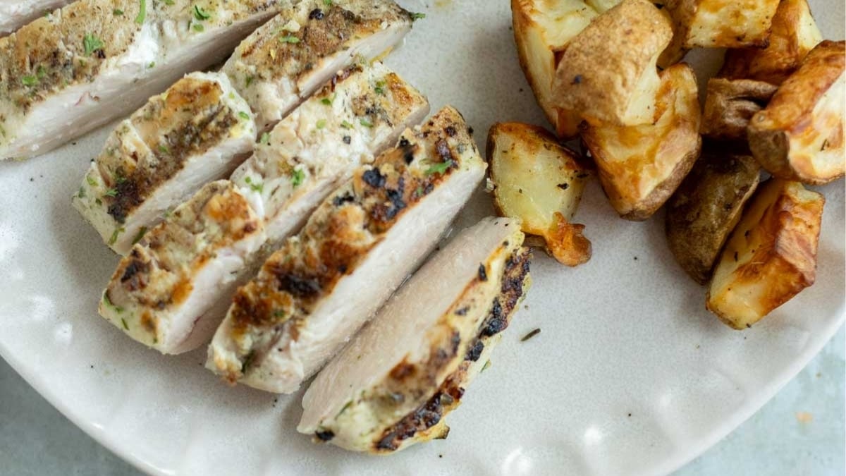 Sliced grilled chicken breast with herbs served next to roasted potato pieces on a white plate.