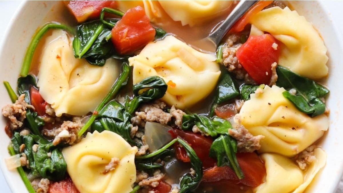 A bowl of tortellini soup with ground meat, spinach, diced tomatoes, and broth, with a spoon partially visible.